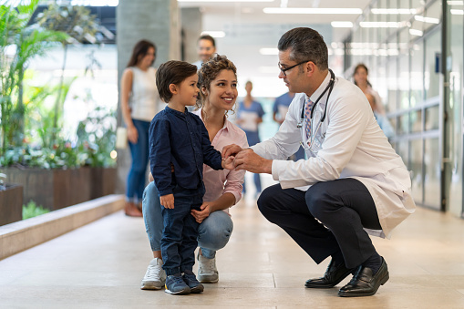 Male pediatrician talking to his little patient who is standing next to his mom all smiling