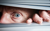 Man's single blue eye peeps fearfully through closed venetian blinds