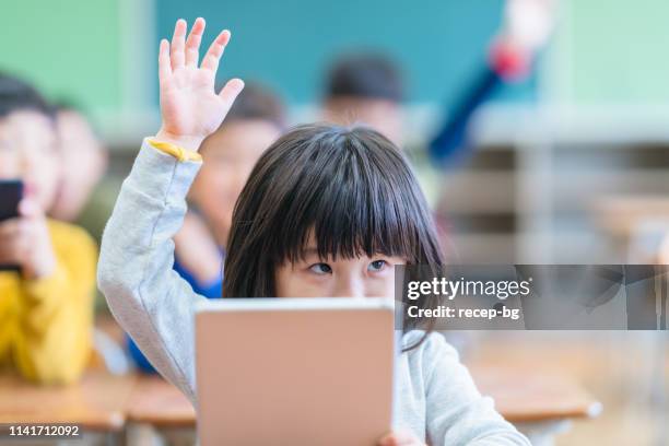 little cute girl holding digital tablet and raising hand in classroom - asian students classroom computer stock pictures, royalty-free photos & images