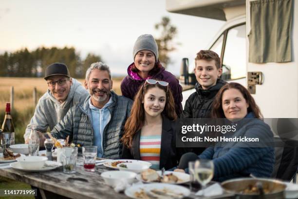 family and friends smile to the camera around a wooden dining table outside a camper van - summer camping new zealand stock pictures, royalty-free photos & images