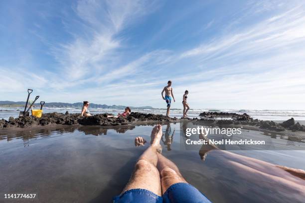 pov shot of a man's legs sitting in the warm spring water of kawhia beach in new zealand, looking out onto their family - personal perspective or pov stockfoto's en -beelden