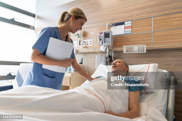 latin american beautiful doctor checking on senior patient lying down on hospital bed talking to him both smiling - doctor looking down stock pictures, royalty-free photos & images