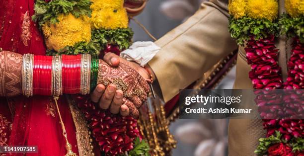 Indian Hindu couple wearing traditional Indian dress and floral garland, holding each other hands during their marriage promising blissful life. Beautiful Henna Mehndi art and bangles on birde hands