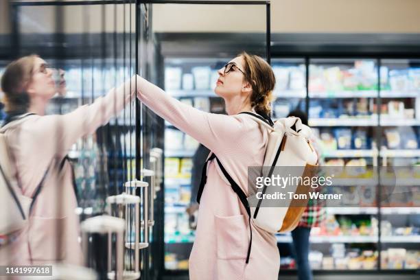 young woman looking in refrigerator at supermarket - supermarket refrigeration stock-fotos und bilder