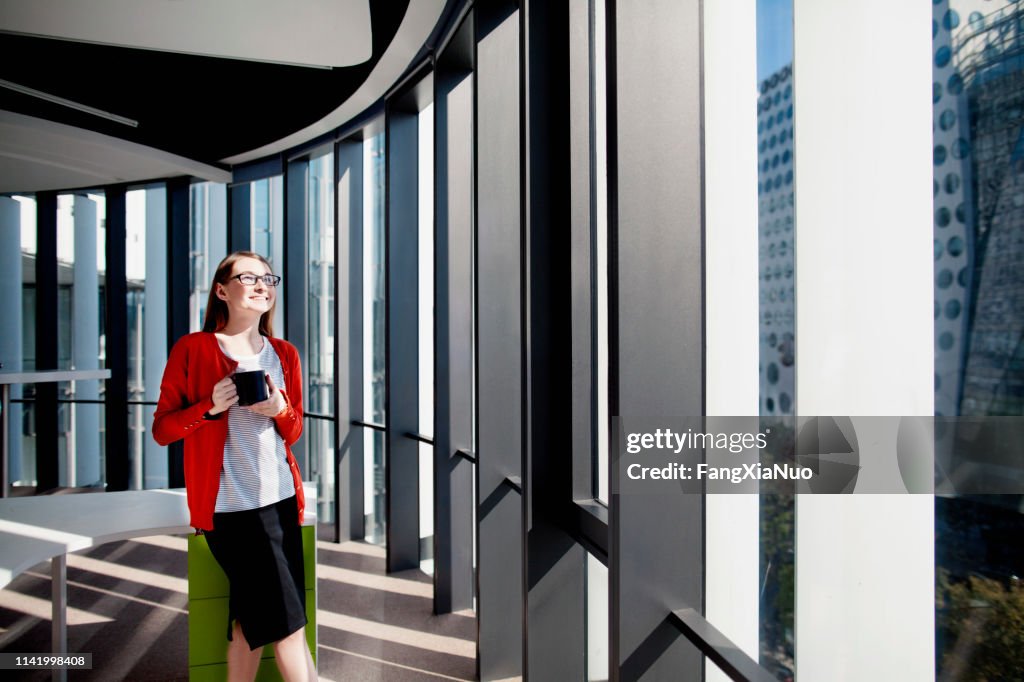 Woman in office smiling in sunshine in window drinking coffee