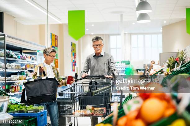 senior couple pushing cart in supermarket - paar gruppierung stock-fotos und bilder