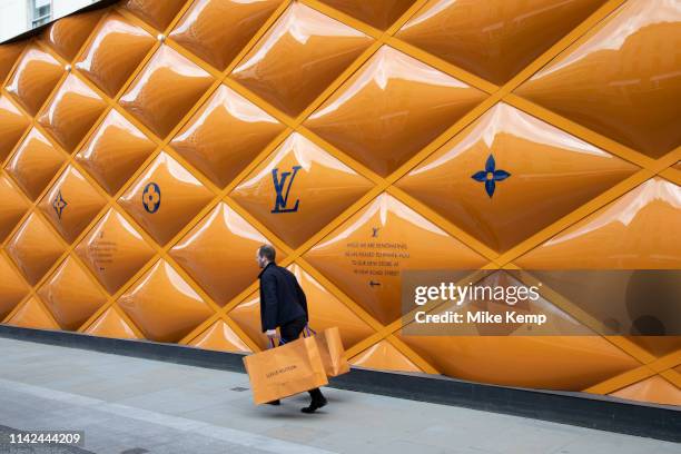 Man with his matching shopping bags passes the yellow temporary hoarding covering exclusive clothes shop window Louis Vuitton on New Bond Street in...