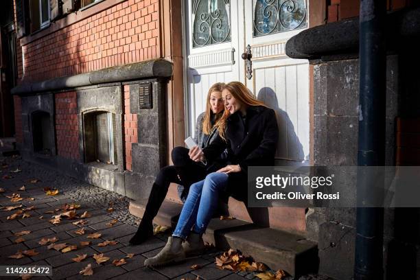 Two playful young women sitting on doorstep using phone