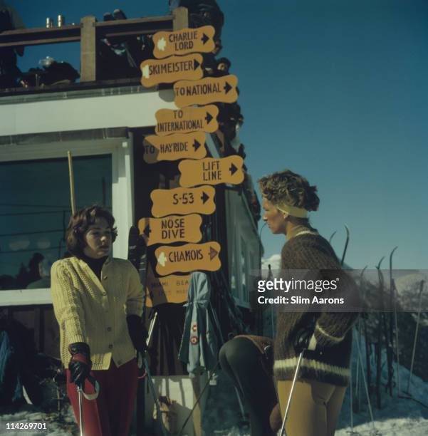 Two women in knitwear, leaning on ski sticks, while standing in the Stowe Mountain ski resort in Stowe, Vermont, 1962. Behind the two women is a sign...