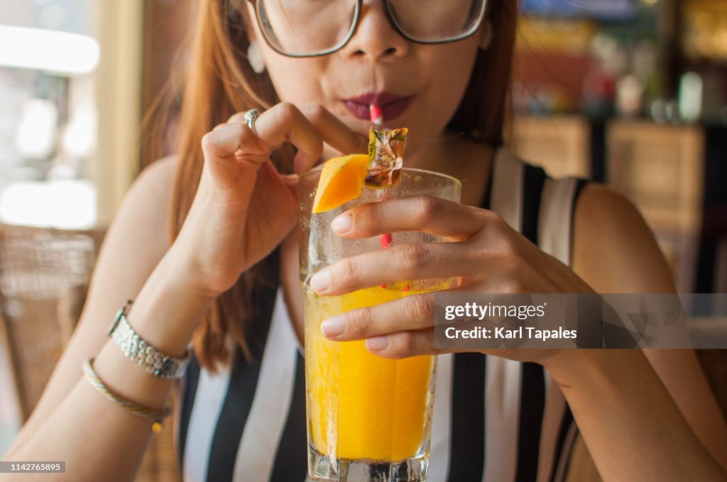 A young woman is drinking a glass of pineapple mango juice with a paper straw