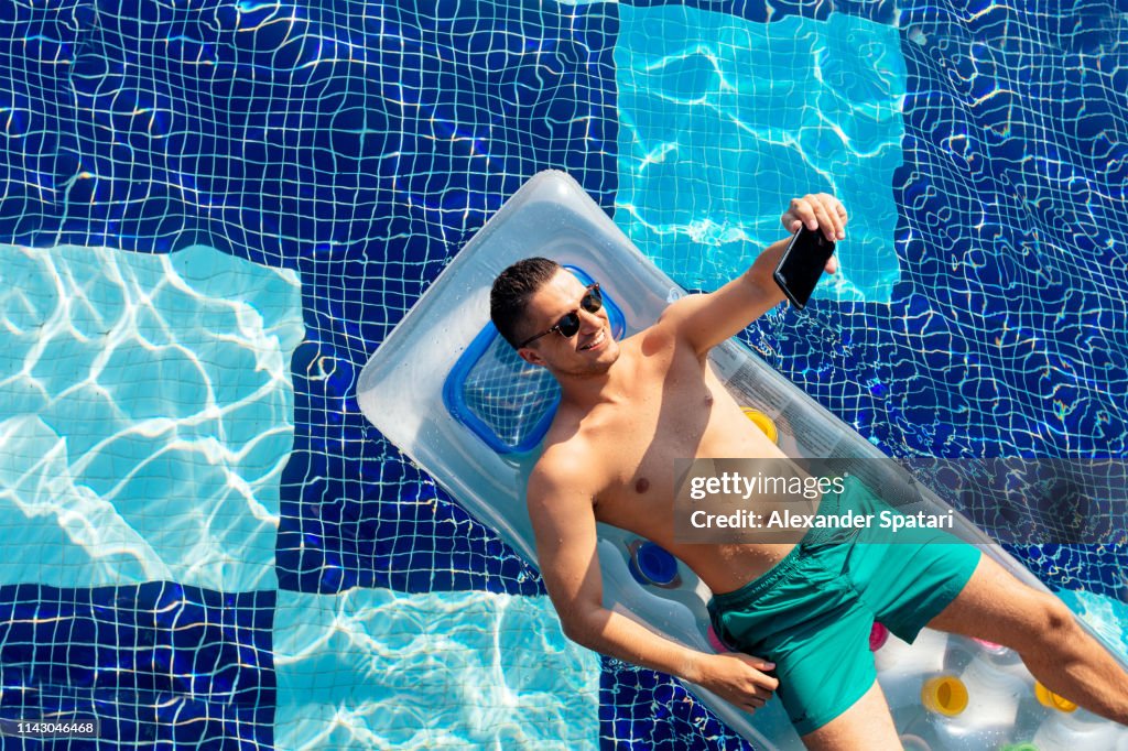 Smiling man in sunglasses on inflatable pool raft taking selfie with smart phone