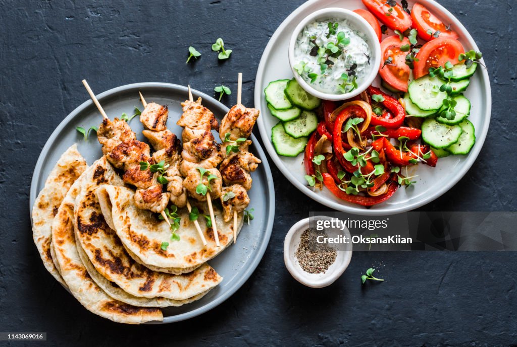 Mediterranean style lunch table - turkey skewers, flatbread, tomatoes, cucumber salad, baked sweet pepper, yogurt herb sauce on a dark background, top view