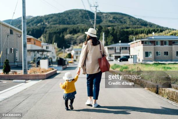 mother and daughter with straw hat holding hands walking along town in the countryside on a sunny day - scenario stock-fotos und bilder