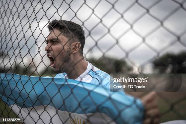 man hanging on fence at sportsfield with argentinian flag - argentina football stock pictures, royalty-free photos & images
