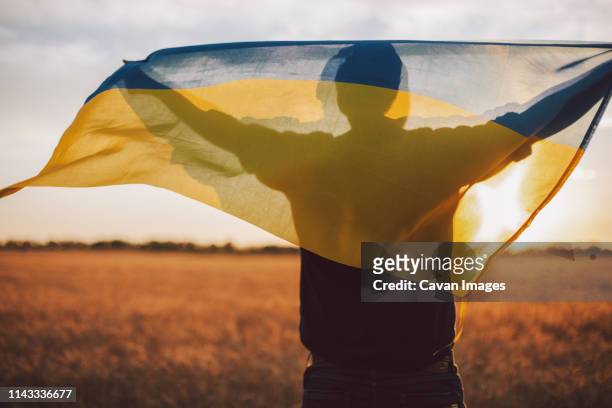 rear view of man with ukrainian flag standing on field during sunset - ukraine war stock-fotos und bilder
