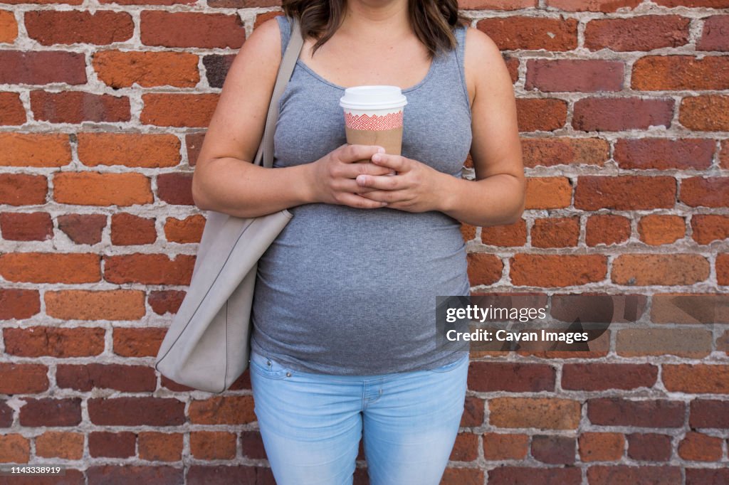 Midsection of pregnant woman holding coffee cup while standing against brick wall