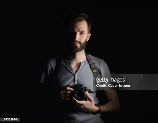 portrait of confident man holding camera while standing against black background - in front of camera stock pictures, royalty-free photos & images