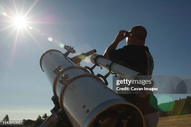 low angle view of astronomer looking at bright sun while standing by telescope against clear sky during sunny day - telescope stock pictures, royalty-free photos & images