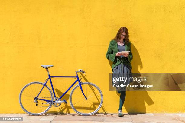 businesswoman using tablet computer while standing on sidewalk by bicycle against yellow wall during sunny day - bike ipad stock pictures, royalty-free photos & images