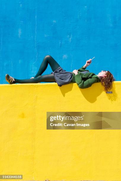 side view of businesswoman using smart phone while lying on yellow wall during sunny day - supino foto e immagini stock