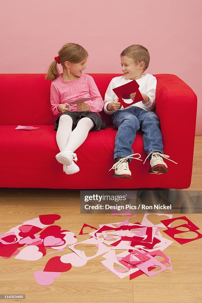A Girl And Boy Sit On A Couch Cutting Out Hearts
