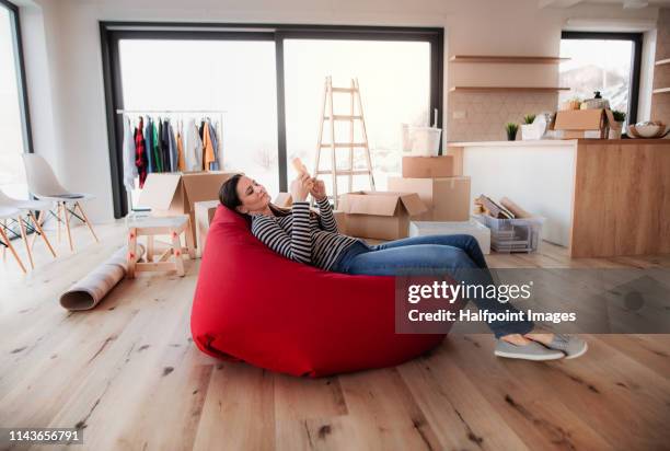 young woman with smartphone sitting on bean bag among moving boxes in a new home. - bean bags fotografías e imágenes de stock