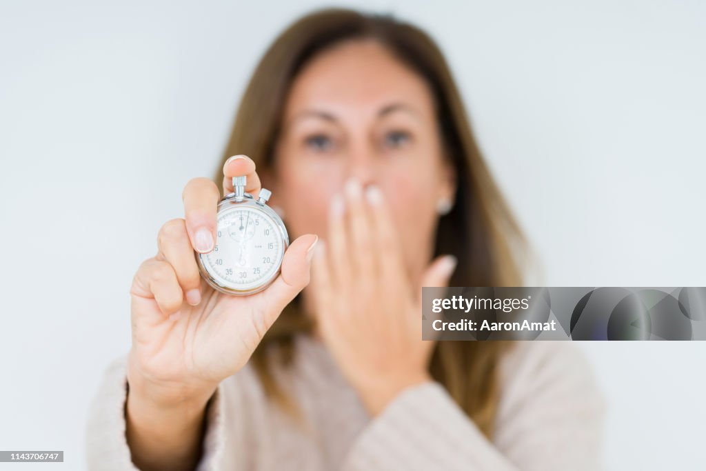 Middle age woman holding stopwatch isolated background cover mouth with hand shocked with shame for mistake, expression of fear, scared in silence, secret concept