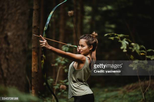 archer fêmea novo que pratica nas madeiras. caça fêmea do caçador na floresta por se durante. - tiro com arco - fotografias e filmes do acervo