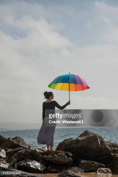 woman holding colorful umbrella on rocky beach, rear view - worry free stock pictures, royalty-free photos & images
