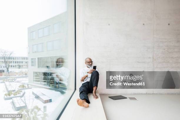 barefoot mature businessman sitting on window sill using smartphone - smart windows stock-fotos und bilder