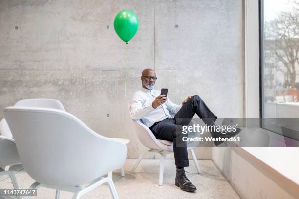mature businessman with green balloon sitting on armchair looking at cell phone - black balloons foto e immagini stock
