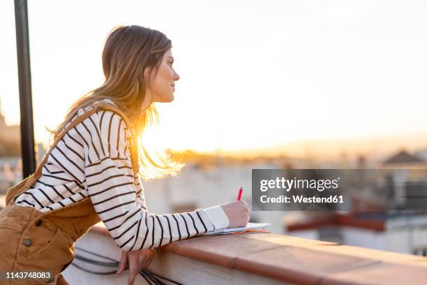 young woman taking notes on roof terrace at sunset - casual woman pensive side view stockfoto's en -beelden