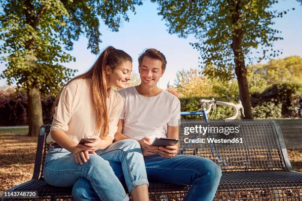 young couple in park sitting on a bench with cell phone and tablet - tablet paar sommer stock-fotos und bilder