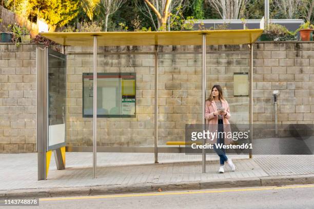 young woman with cell phone waiting at bus stop - aspettare foto e immagini stock