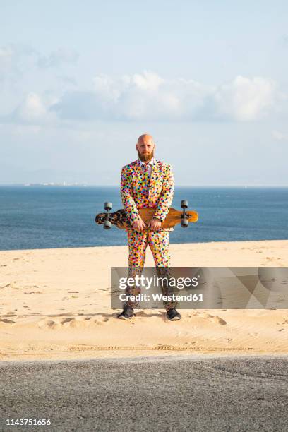 bearded man with longboard wearing suit with colourful polka-dots standing on sand dune - flerfärgad kostym bildbanksfoton och bilder