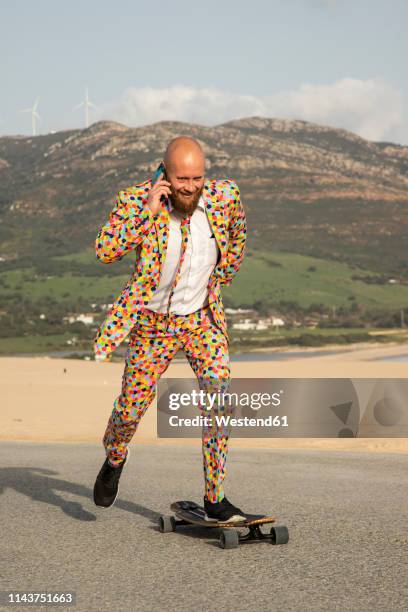 smiling bald man on the phone wearing colourful suit while skateboarding on road - single lane road - fotografias e filmes do acervo