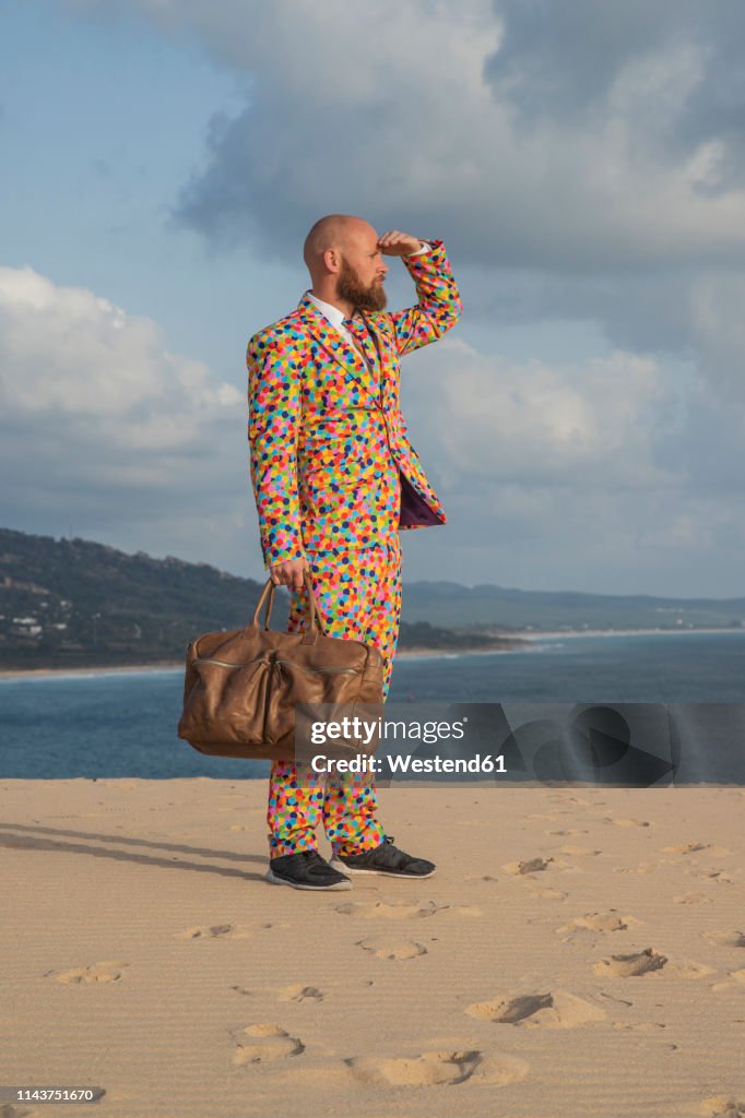 Spain, Tarifa, man with travelling bag wearing suit with colourful polka-dots standing on dune looking at distance