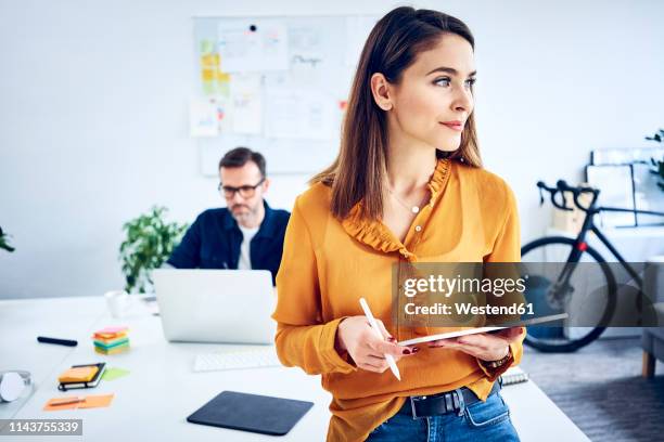 businesswoman holding tablet in office with colleague in background - graphics tablet stock pictures, royalty-free photos & images
