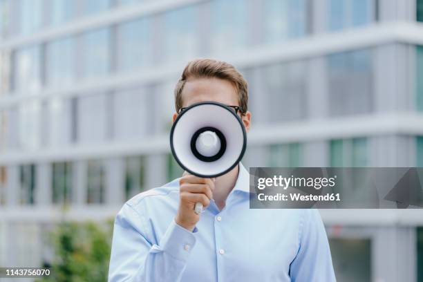 young businessman standing in front of modern office building, using megaphone - megafon stock-fotos und bilder