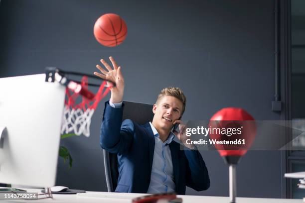 young businessman sitting at desk in office playing basketball and talking on cell phone - basketball all access stock pictures, royalty-free photos & images