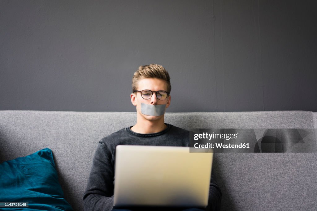 Young man with taped mouth sitting on couch using laptop