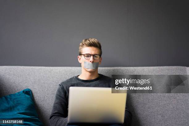 young man with taped mouth sitting on couch using laptop - censorship imagens e fotografias de stock