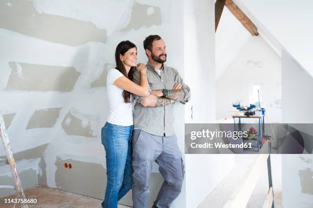 smiling couple in attic to be renovated looking out of window - house renovation stock pictures, royalty-free photos & images