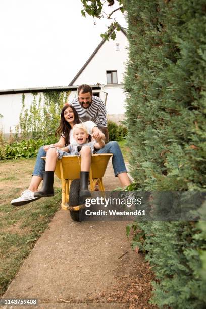 playful man pushing wife and son sitting in wheelbarrow in garden - schubkarre stock-fotos und bilder