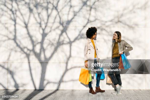 two happy women with shopping bags standing at a wall talking - two women talking stockfoto's en -beelden