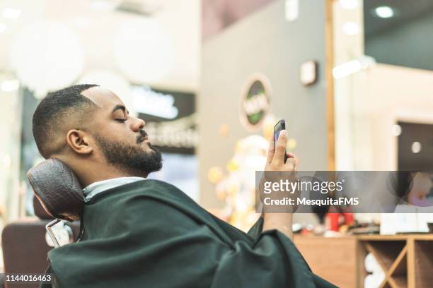 african man using cell phone in barbershop - african american hair salon stock pictures, royalty-free photos & images
