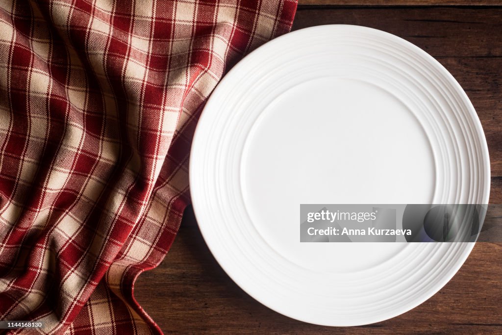 Empty white plate with a napkin on an old wooden brown background, top view. Image with copy space. Kitchen table with a towel and a plate - top view with copy space.