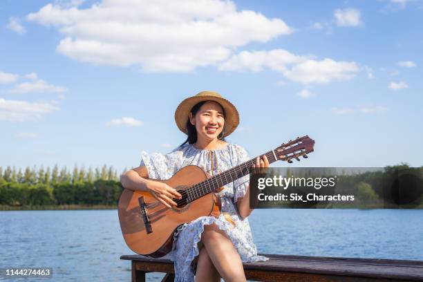 happy young asian woman playing classical guitar at lakeside and blue sky in background - country and western music stock pictures, royalty-free photos & images