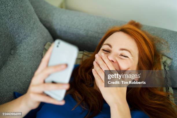 woman lying on sofa using smart phone, smiling behind hand - reírse mujer fotografías e imágenes de stock