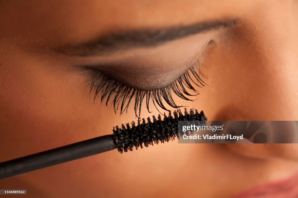 Closeup of a young dark-skinned woman applying mascara on a white background